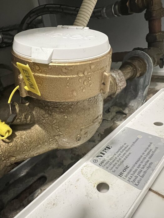 Vattenmätare med kondens, rör och installationsinstruktioner synliga. Inomhus, närbild, teknisk utrustning.