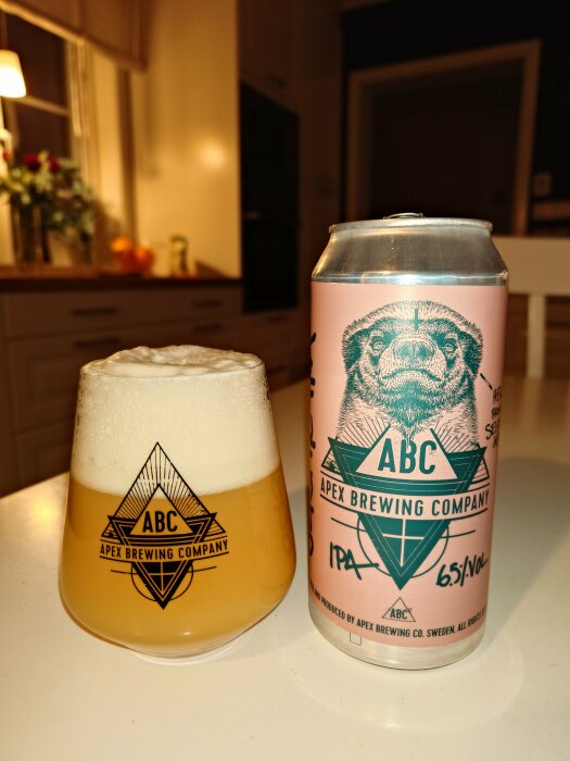 Ölglas med skum vid sidan av ölburk. Apex Brewing Company logotyp. Inomhus, köksmiljö i bakgrunden.