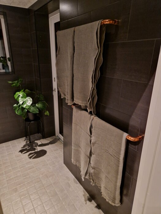 Mörkt badrum med handdukar på kopparstång, krukväxt på pall, kaklade väggar och golv.