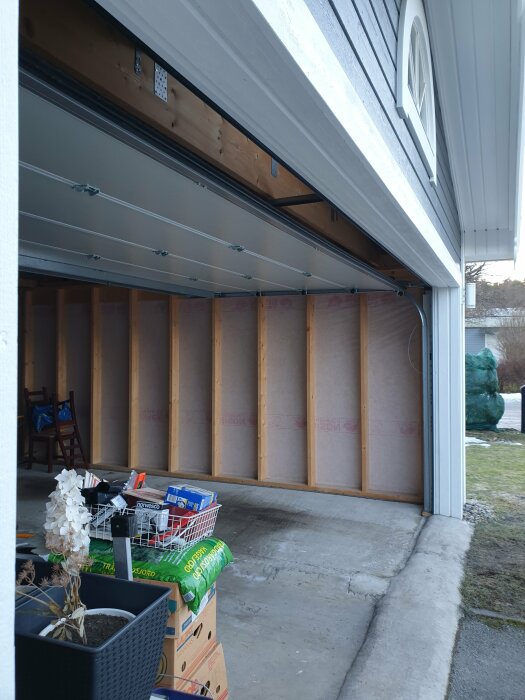 Öppen garageport visar ett tomt garage med isolering, några lådor och krukväxter i förgrunden.