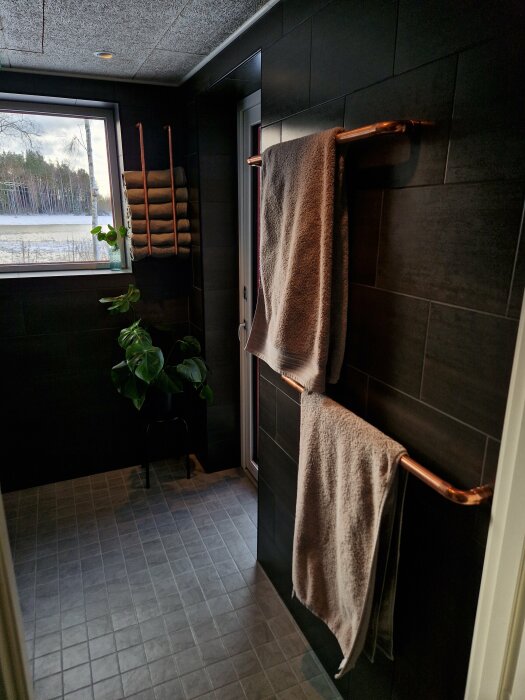 Modernt badrum med kopparhandduksstänger, gröna växter och utsikt över vinterlandskap genom fönstret.