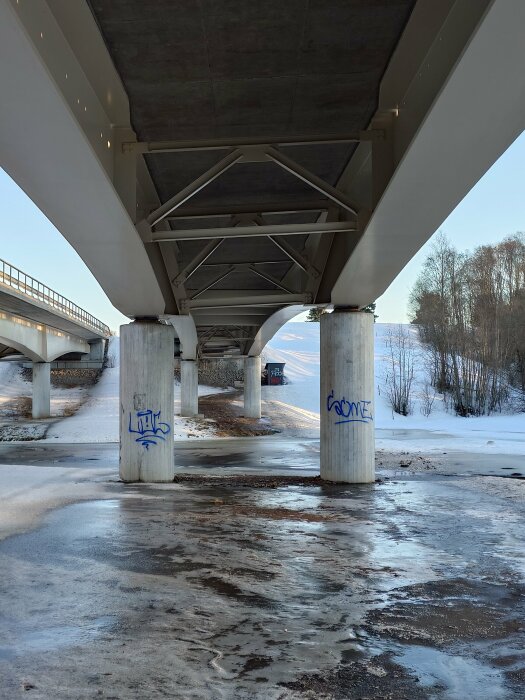 Brokonstruktion underifrån, betongpelare, snö, is, graffiti, vinterdag, träd i bakgrunden.