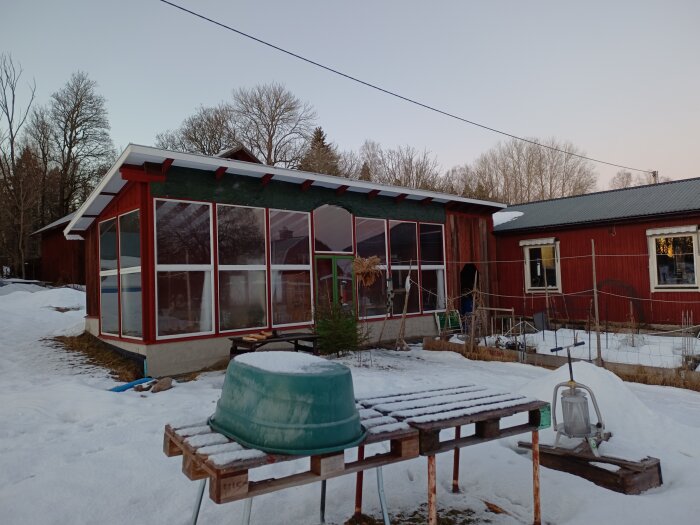 Ett växthus med rött tak mot en snöig trädgård, röda byggnader, och diverse trädgårdsredskap i förgrunden.