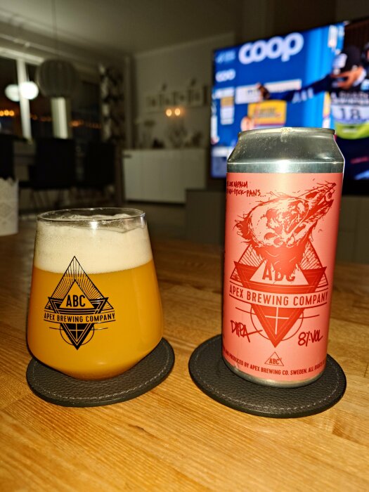 Ölglas med skum, burk från Apex Brewing Company, inomhusmiljö med TV i bakgrunden.