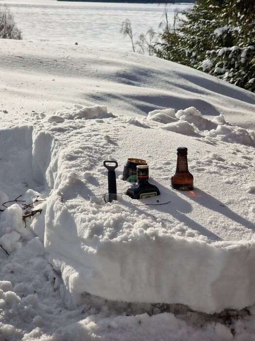 En flaska, burk och öppnare på ett snötäckt bord utomhus med skogsbakgrund och solsken.