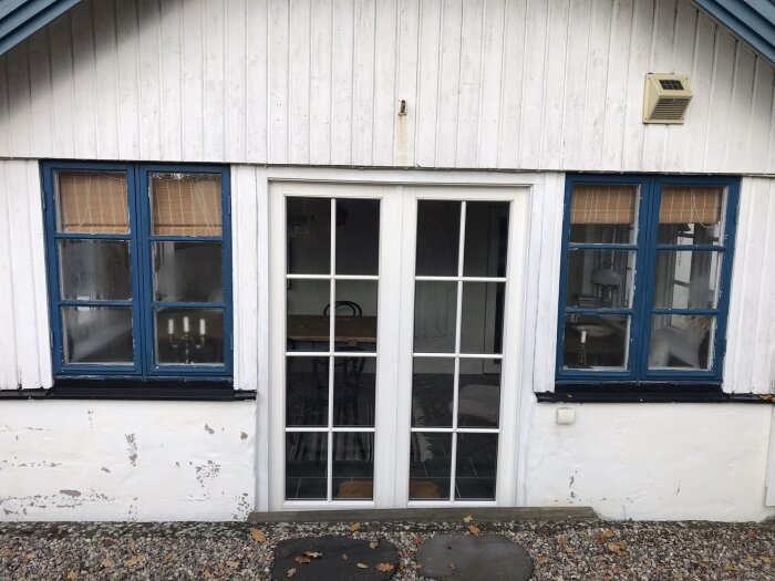 Vit husfasad med blå fönsterluckor och en vit dubbeldörr med glaspaneler. Luftkonditioneringsenhet ovanför till höger.