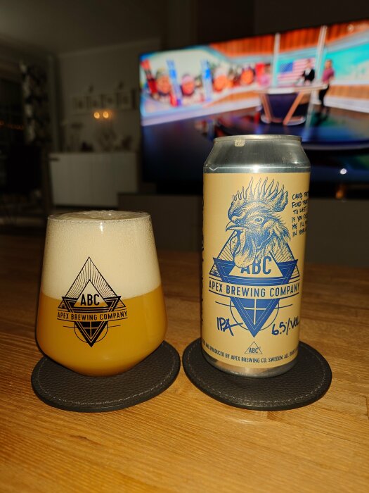 Ölglas och burk från Apex Brewing Company framför suddig TV. Skumtopp på öl. Hembarambians.