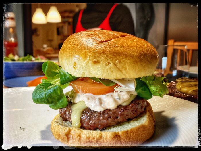 Hamburgare med sallad, tomat, ost och sås, inomhus, en person i förkläde i bakgrunden, mysig matsituation.