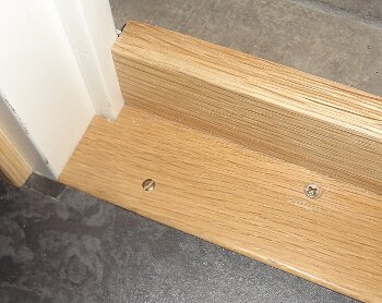 Tröskel av trä vid en dörröppning med skruvar installerad på ett grått golv.