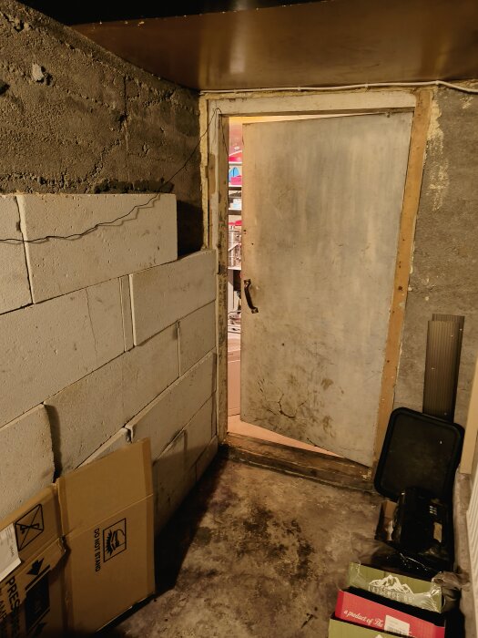 Källarrum med betongväggar, grov dörr, lådor, skräp, synlig röran genom dörrspringan.