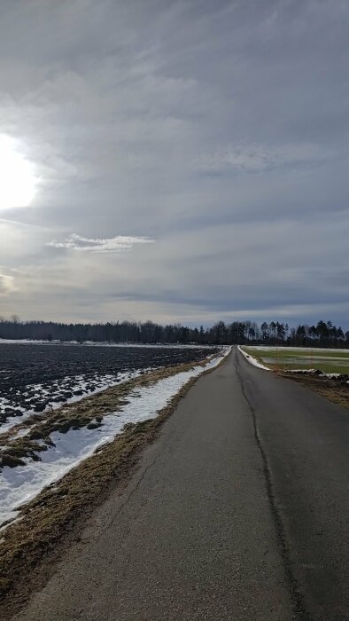 Lantväg, delvis snötäckt fält, moln, bar himmel, träd i horisont, vinter till vårig känsla.