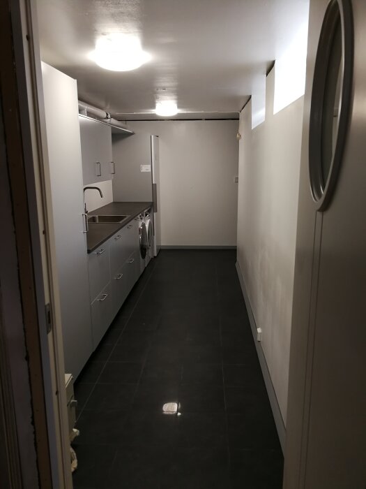 Lång korridor med tvättmaskin, torktumlare, skåp och bänk, belysning i tak, mörkt golv.