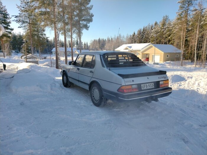 Klassisk bil, Saab 900, parkerad i snöigt landskap, soligt vinterdag, hus och träd i bakgrunden.