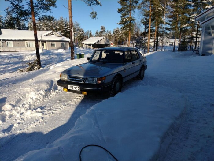 Gammal bil parkerad framför huset, snötäckt landskap, vinter, soligt, träd, stillhet, kallt.