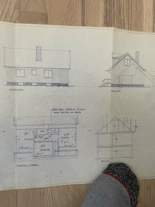 Arkitekturritningar av hus, planlösning och fasader, på öppen bok, personens fot syns nederkant.