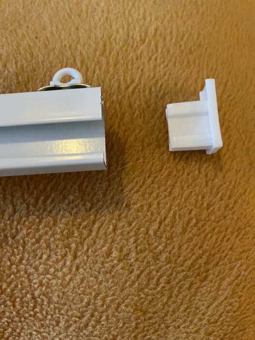 Två vita plastdelar på brun matta; ser ut som delar från gardinupphängning eller persienn.