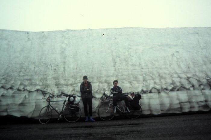 Två personer med cyklar framför stor snövägg, grå himmel, äventyr, kyligt väder, retrokläder.