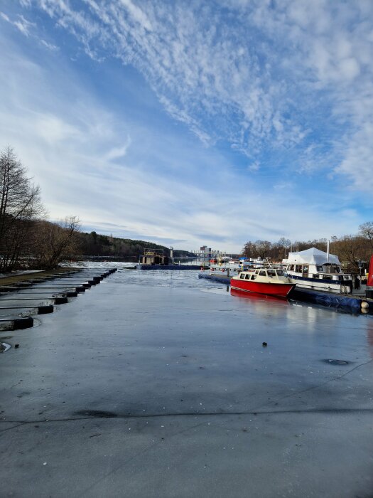 Istäckt kanal med båtar, träd, delvis molnig himmel, stadssiluett i bakgrunden.