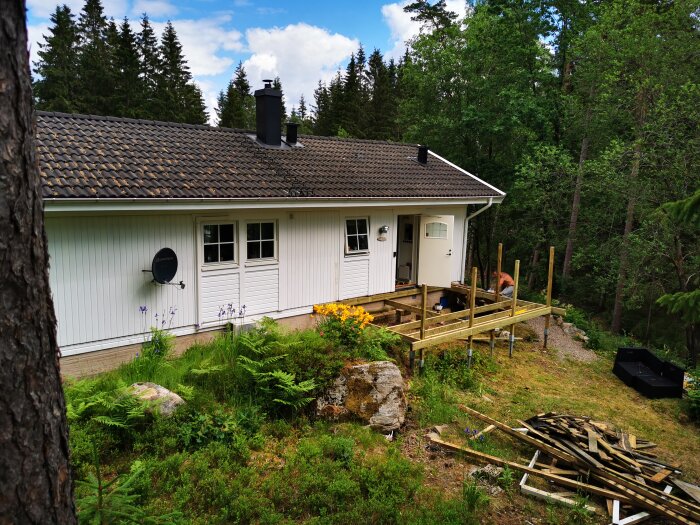 Ett enfamiljshus med vit fasad, svart tak, i skog. Byggnation av trädäck pågår. Lugnt, lantligt boende.