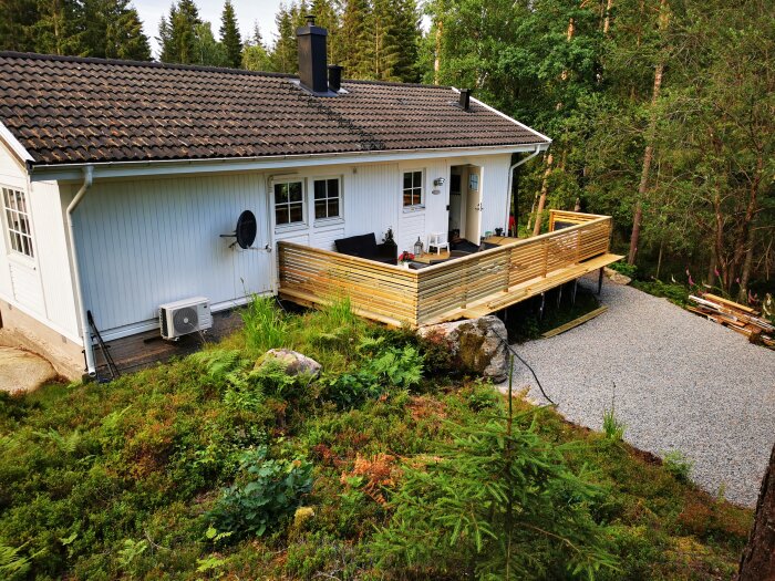 Ett vitt hus med veranda och skog runtomkring, grusgång, grönska, luftvärmepump och uteplats.