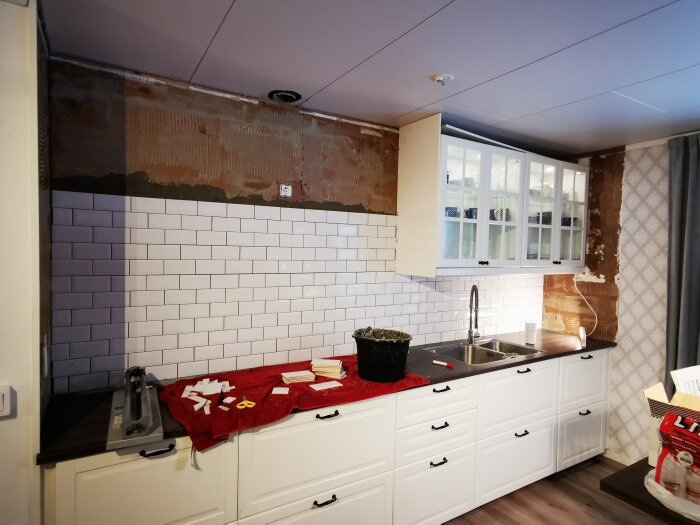 Kök under renovering, vit kakelvägg halvfärdig, skåpluckor och diskho, byggmaterial och verktyg synliga.