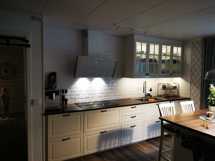 Modernt kök med vit kakelvägg, köksö, inbyggd belysning, och en person i bakgrunden.