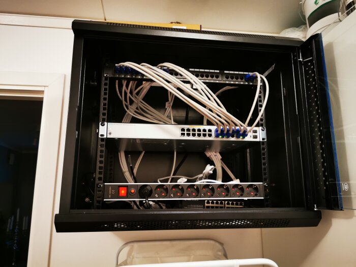 Svart nätverksskåp med patchpaneler, kablar och strömfördelningsenhet monterade i en vägg.