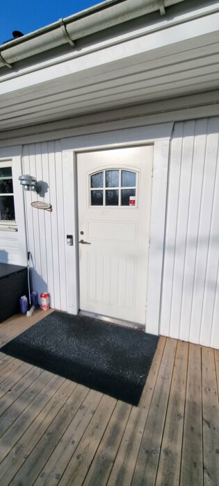 En vit ytterdörr med fönster och lås på ett hus, trädäck, matta, och utebelysning under dagtid.
