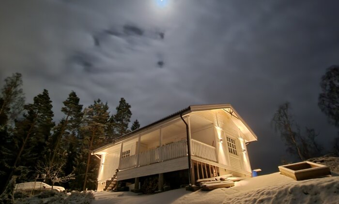Ett upplyst hus på pålar i snölandskap nattetid med moln och månsken över trädtopparna.