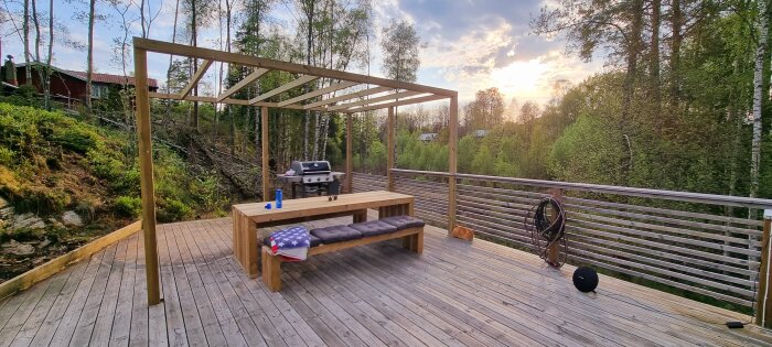 Trädäck med pergola, bänkbord, grill, skogsbakgrund, solnedgång, klart väder, avslappnad utomhusmiljö.