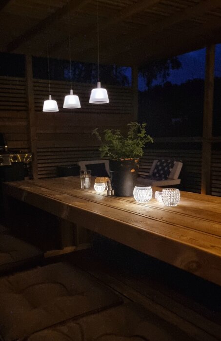 Kväll, belyst veranda, träbord, lampor, glas, krukväxt, mysig atmosfär.