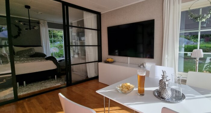 Modernt vardagsrum med glasvägg till sovrum, TV, matbord, öl och snacks.