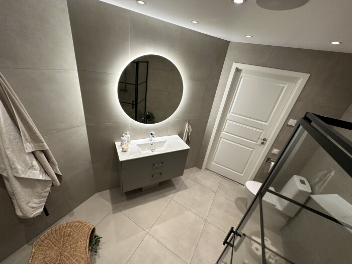 Modernt badrum, spegel med belysning, duschhörn, flytande handfat, grå kakel, vita detaljer, badrock, korgstol.