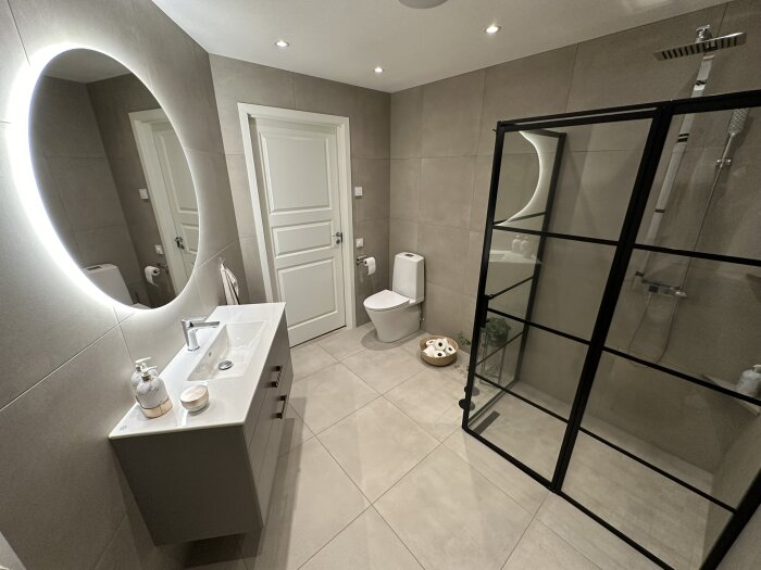 Modernt badrum med duschhörna, toalett, tvättställ, spegel, och gråa kakelväggar.
