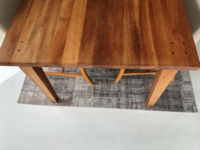 Ett träbord med synliga skruvhål, över en texturerad, grå matta på ett ljust golv.