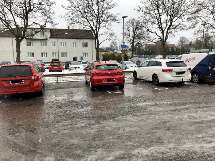 En parkeringsplats med olika bilar, våta ytor, lite snö, träna och byggnader i bakgrunden.