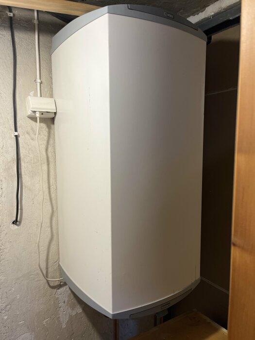 Vit varmvattenberedare monterad på en vägg i ett trångt utrymme med synliga rör och kablar.