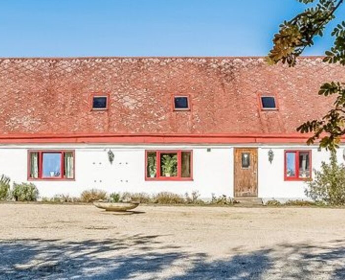 Ett vitt lantligt hus med röd tegeltak, vindsvåningsfönster och röda fönsterkarmar, under en klarblå himmel.