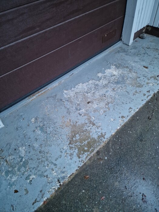 Garageingång med slitna betonggolv och fläckar, brun garageport, en grå sten i hörnet.