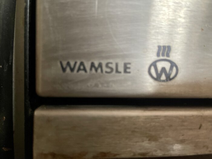 Närbild på metallisk yta med texten "WAMSLER" och stiliserad logotyp.