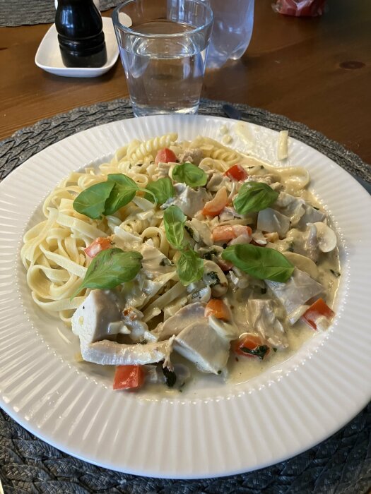 Pasta med krämig sås, kyckling, grönsaker och färsk basilika serveras på en tallrik med vattenglas i bakgrunden.