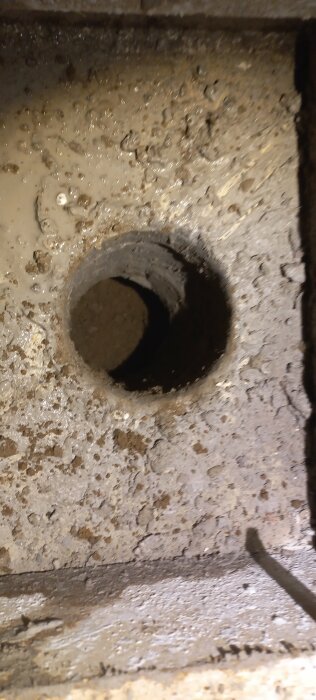 Närbild av ett smutsigt avloppshål i betong med fukt och partiklar runt kanterna.