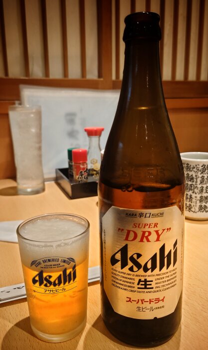 Asahi ölflaska och glas på bord, japansk restaurangmiljö, dryck serverad, bordskondiment i bakgrunden.