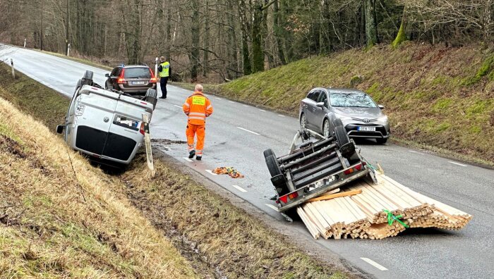 Trafikolycka, bil på sidan, arbetsfordon, träplankor på vägen, personer utvärderar situationen, fuktig asfalt.