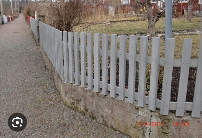 Grått staket längs grusgång, träd, röda hus i bakgrunden, daterad bild.