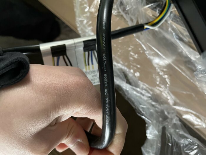 En hand håller en elektrisk kabel med tekniska specifikationer synliga, omgiven av oskarpa föremål och plast.