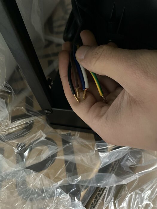 En hand håller flerkärniga ledningar med avskalade ändar framför en svart elektronisk apparat, med plastförpackning synlig.