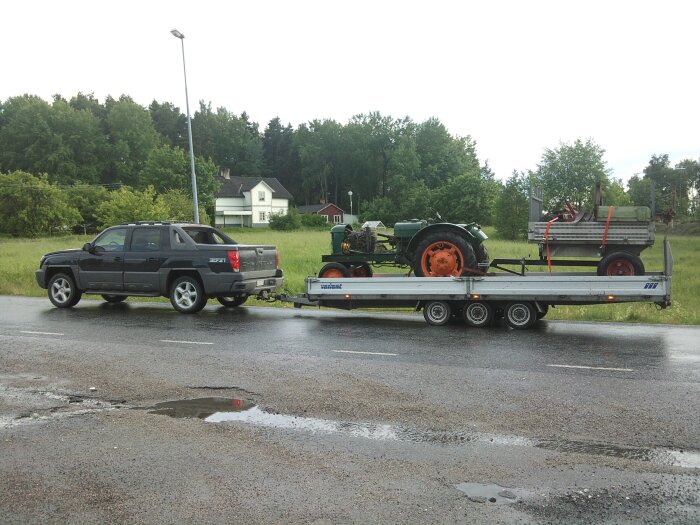 Pickup drar släpvagn med traktor och höbalare, gråmulen dag, våt väg, gröna träd, hus i bakgrunden.