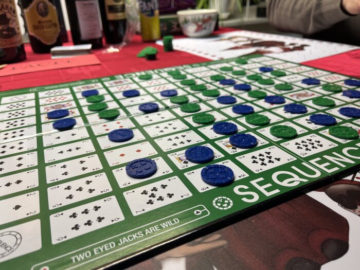 Brädspel "Sequence" i spel, blå och gröna markörer, alkoholflaskor i bakgrunden, sällskapsspel.