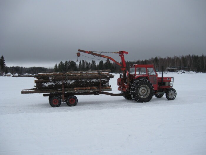 Röd traktor med timmersläp fullt med stockar på snötäckt mark under molnhimmel.